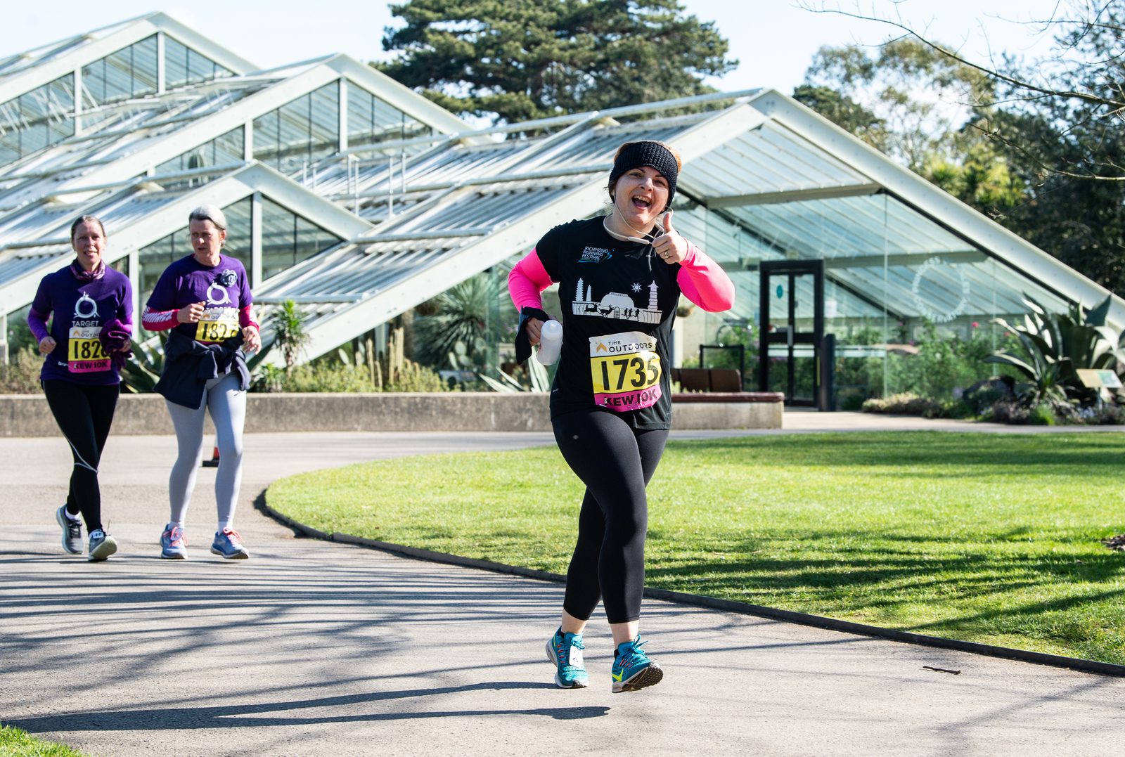 Three women are participating in a run, wearing numbered bibs. The woman in the front, wearing a black outfit with a beanie, is smiling and pointing toward the camera. Two women in purple shirts follow behind her. They are outside with glass-roofed structures in the background.