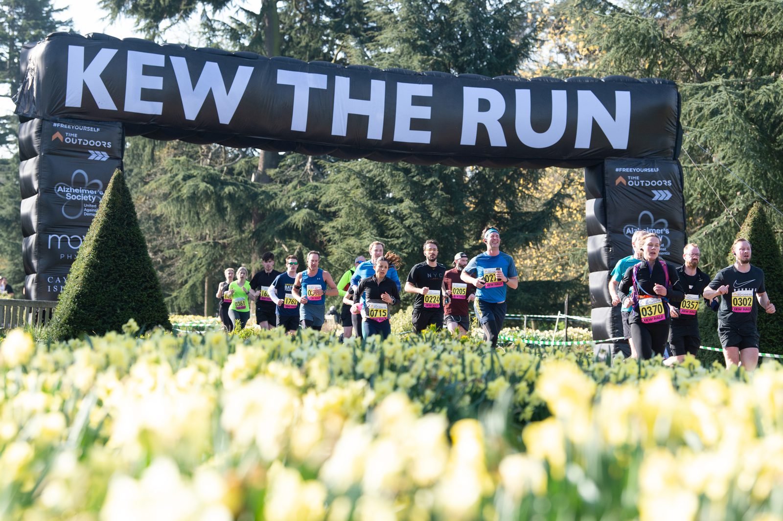 A group of runners participating in a race pass under an inflatable archway that reads "KEW THE RUN." The scene is set in a lush, wooded area with blooming flowers in the foreground. The runners wear race bibs and exercise gear, showing determination and enthusiasm.