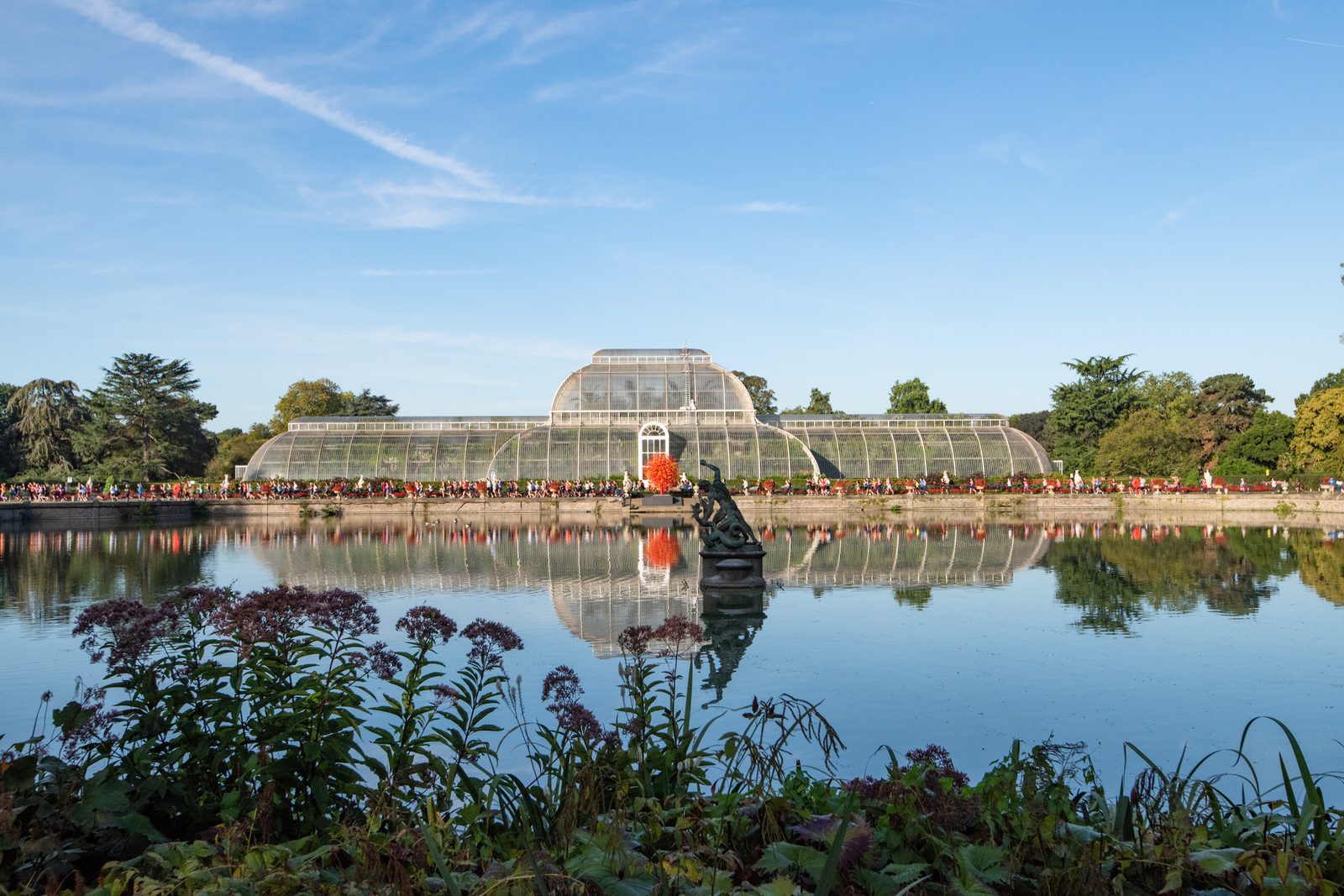 A large glass greenhouse stands symmetrically behind a serene lake, reflecting the structure and the blue sky above. In the foreground, leafy plants and flowers border the water. Trees surround the greenhouse, adding greenery to the picturesque scene.