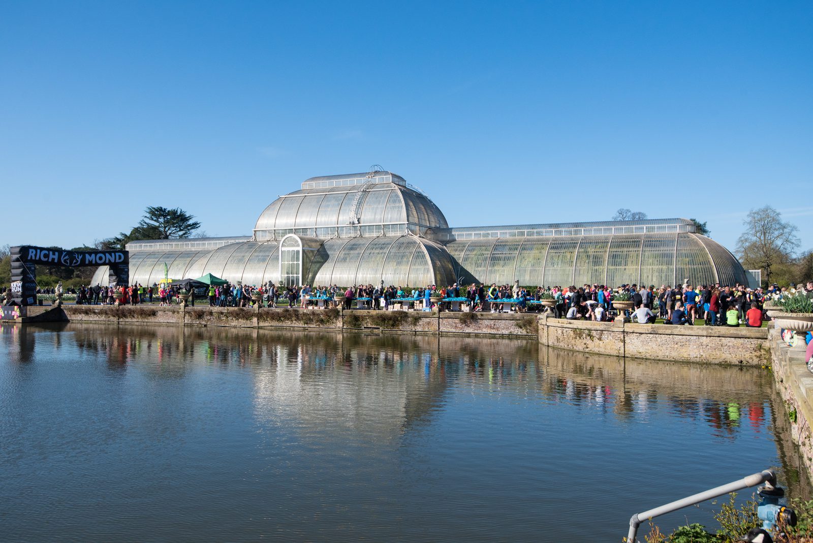 A large crowd gathers outside a historic glasshouse with a pond in the foreground under a clear blue sky. A banner reading "Richmond" is visible to the left.