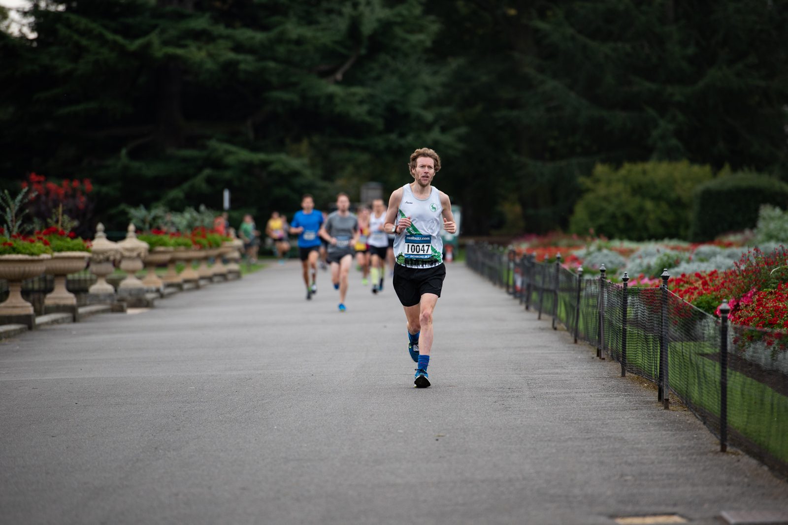 A runner with bib number 10071 leads a group during a race in a park, surrounded by greenery and colorful flowers. He is focused and slightly ahead of several other runners, moving along a paved path lined with benches and flowerbeds.