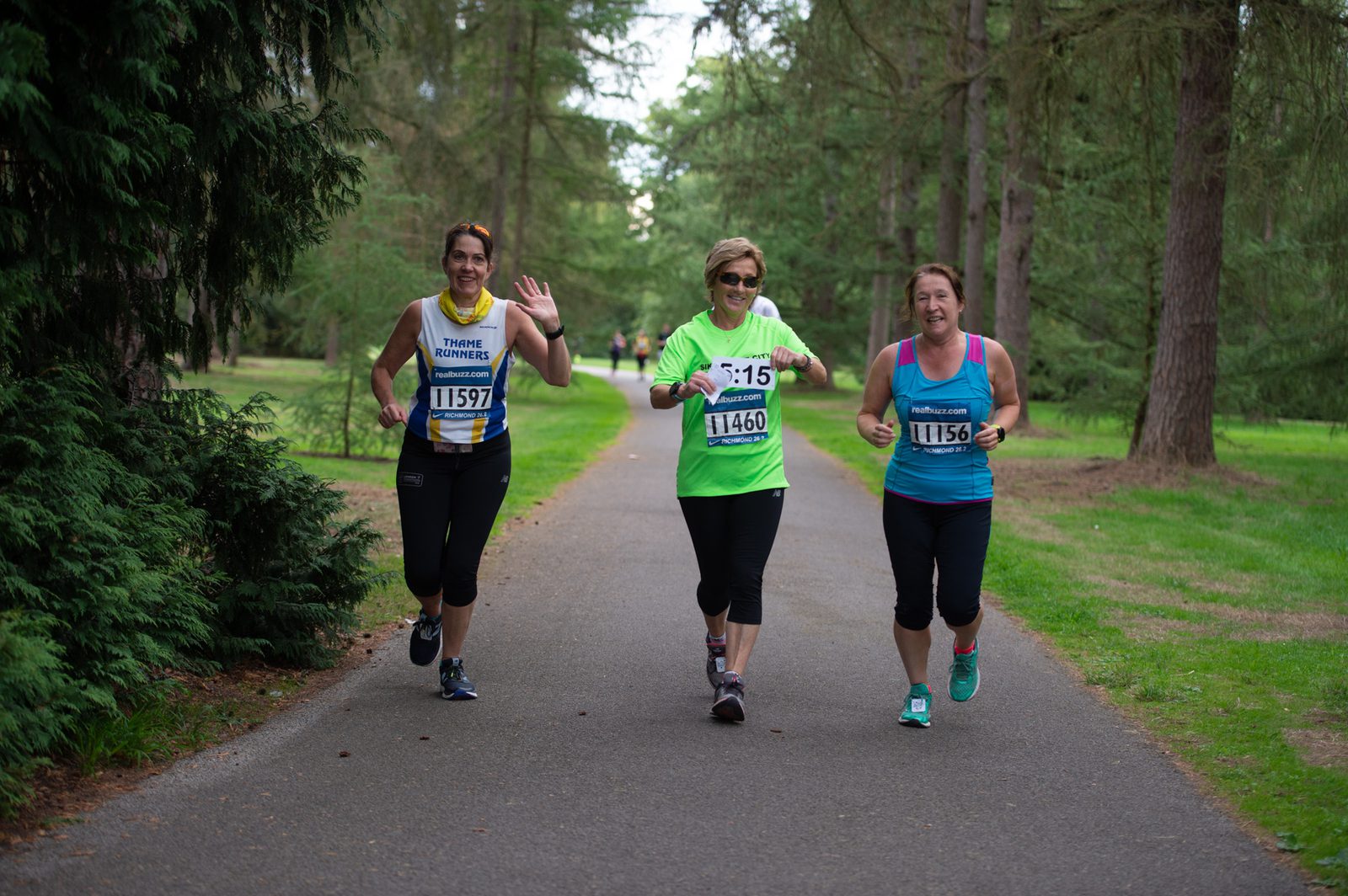 Three women are running on a forest trail during a race. All are smiling, with the woman on the left waving. They are wearing different colored tops (yellow, green, and blue) and have bib numbers pinned to their shirts. The path is surrounded by tall trees and grass.