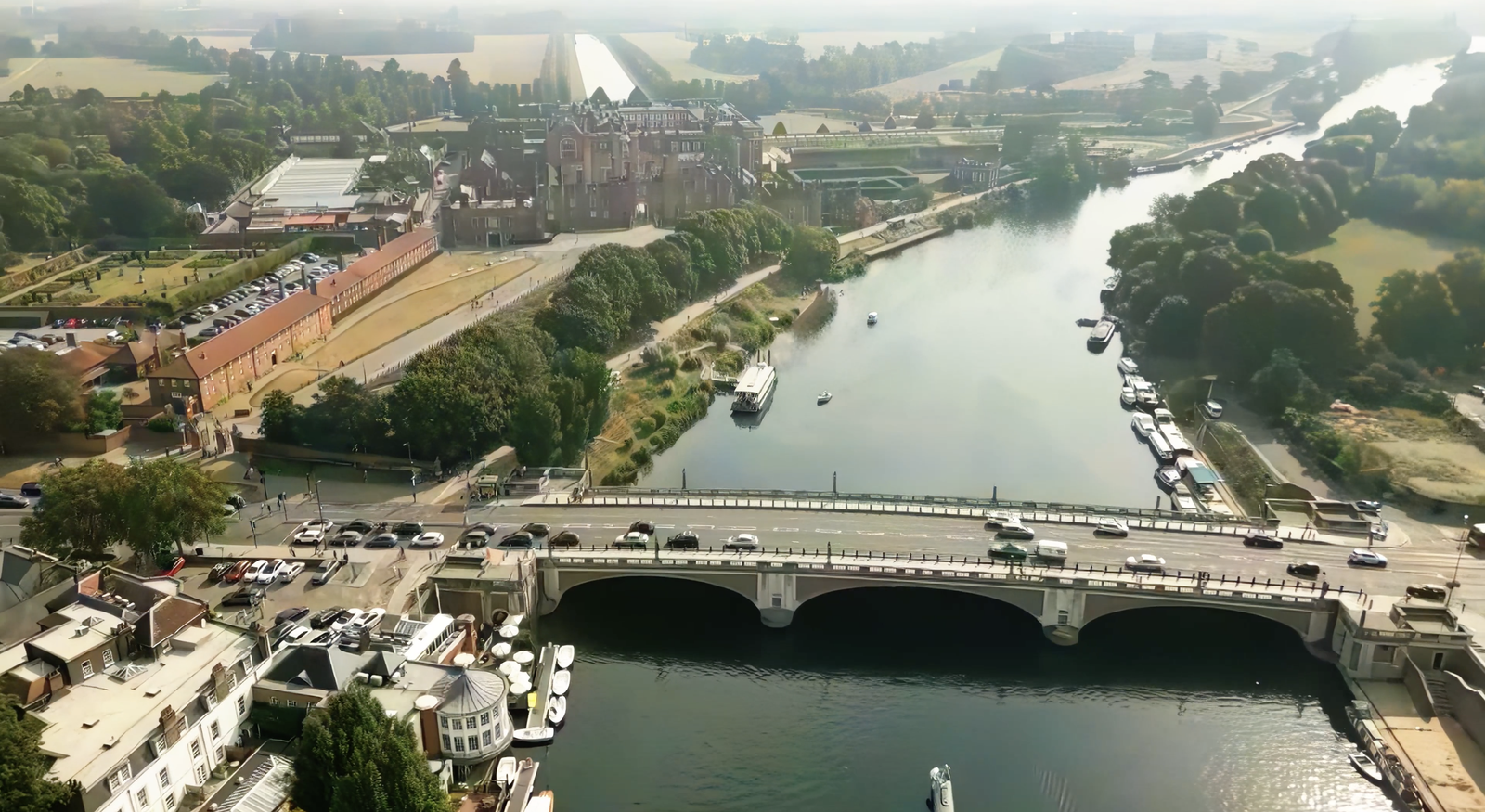 Aerial view of a picturesque river with a bridge spanning across it. The river is lined with trees and boats. In the background, a large, historic brick building sits surrounded by lush greenery under a partially cloudy sky.