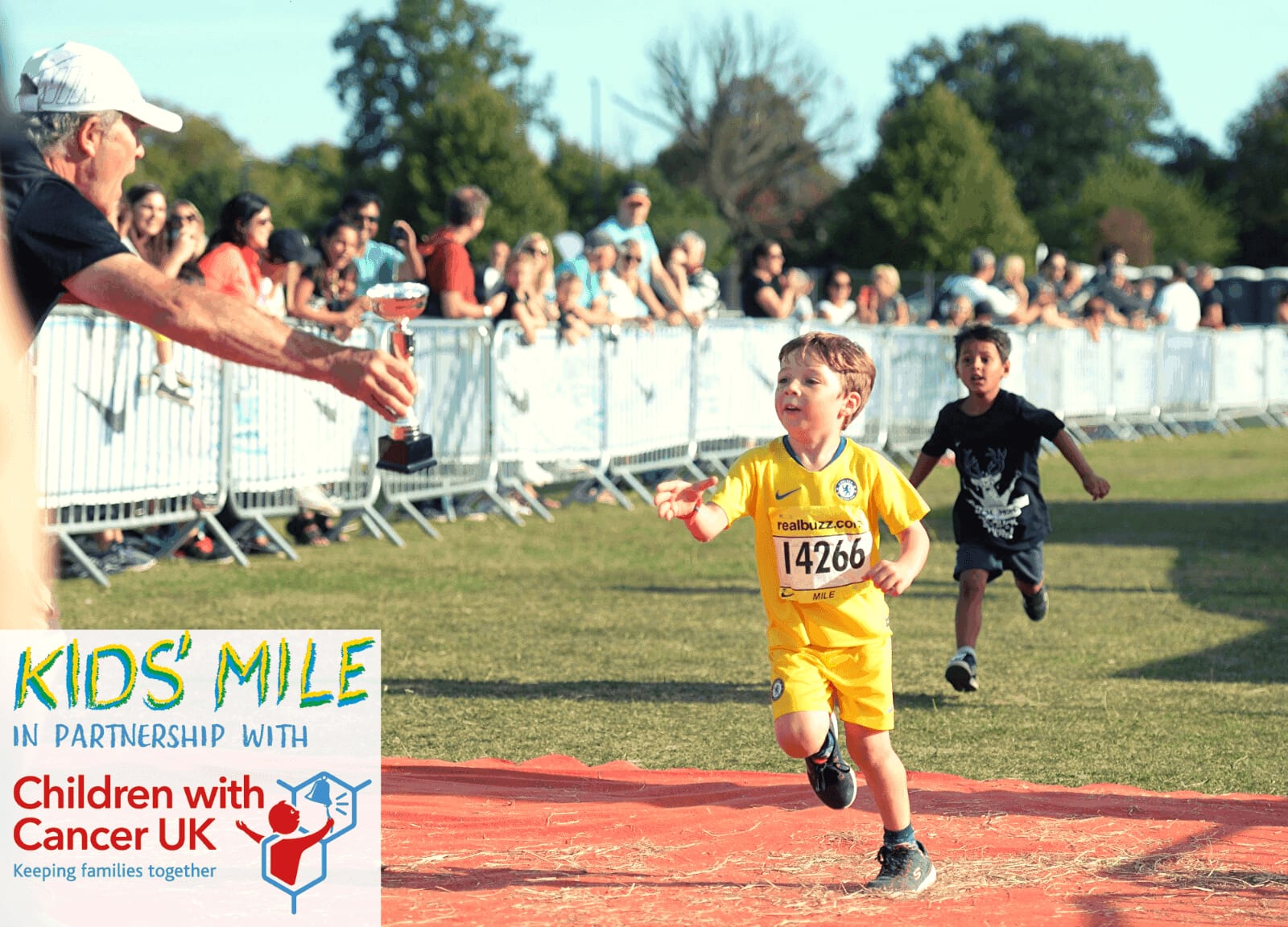 A young child dressed in yellow is running toward the finish line in a race, with an adult handing out water nearby. Another child is running behind him. Spectators are gathered behind barriers, cheering. A banner reads "Kids' Mile in partnership with Children with Cancer UK.