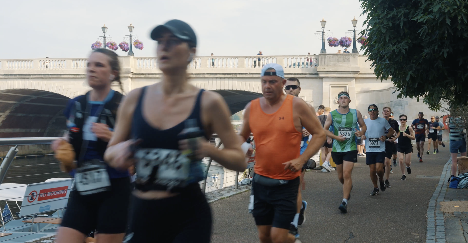A group of runners participates in a race along a riverside path, with a bridge and lamp posts decorated with flowers in the background. The runners are wearing athletic gear, including tank tops, caps, and race bibs, and are focused on the race.