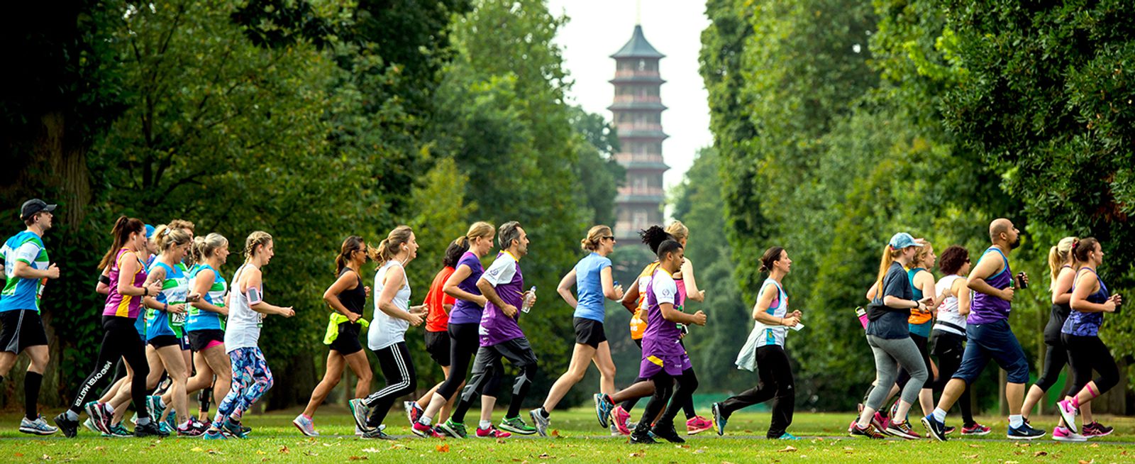 A diverse group of people, dressed in colorful athletic wear, are running through a lush, green park. In the background, a large pagoda structure is visible among the trees. The runners appear to be participating in a group exercise or race.