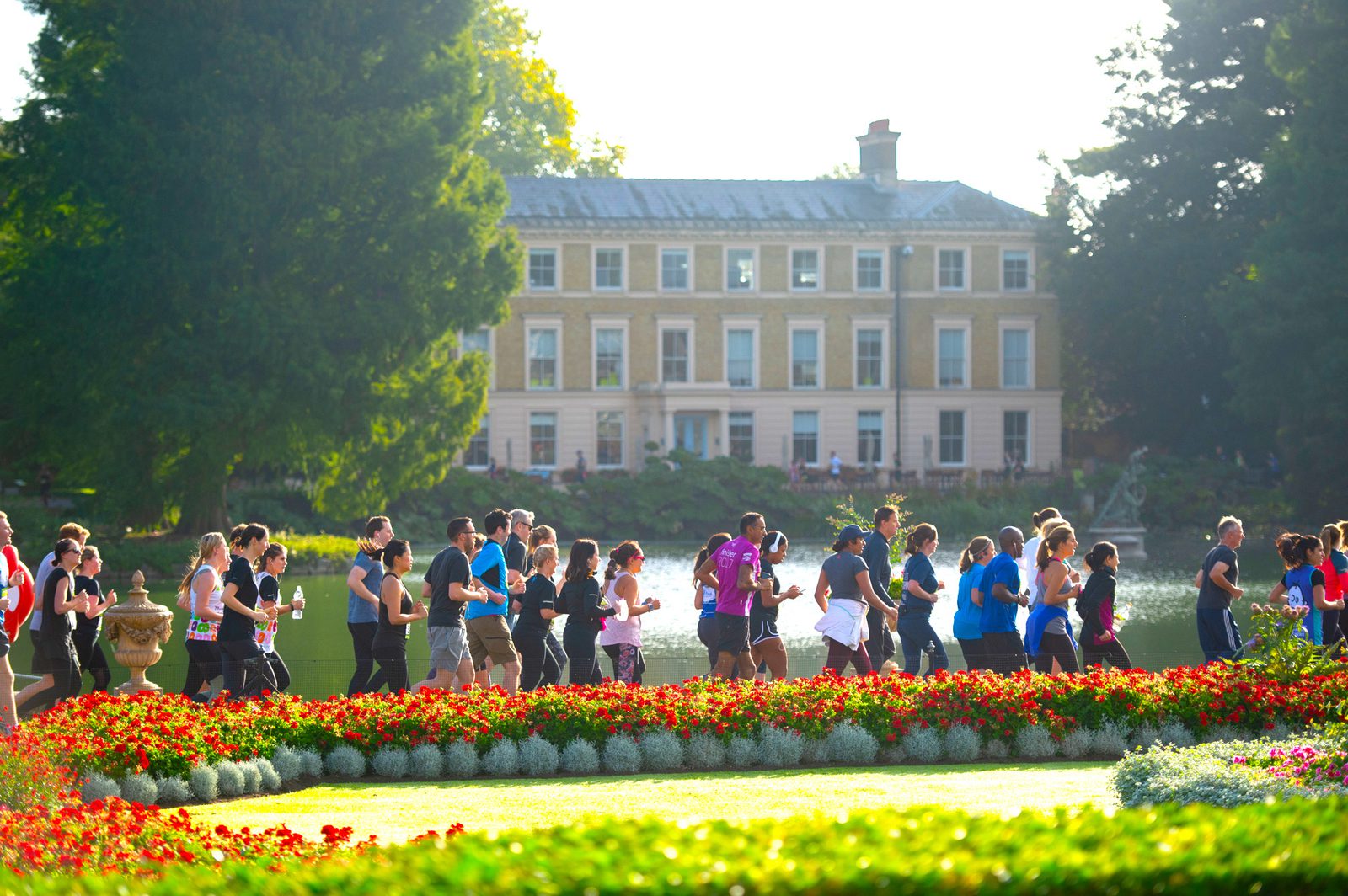A group of people participating in a community run, jogging along a path in a beautifully landscaped park. The backdrop includes a stately mansion and vibrant flowerbeds. The sun is shining, and the greenery is lush, creating a picturesque setting.