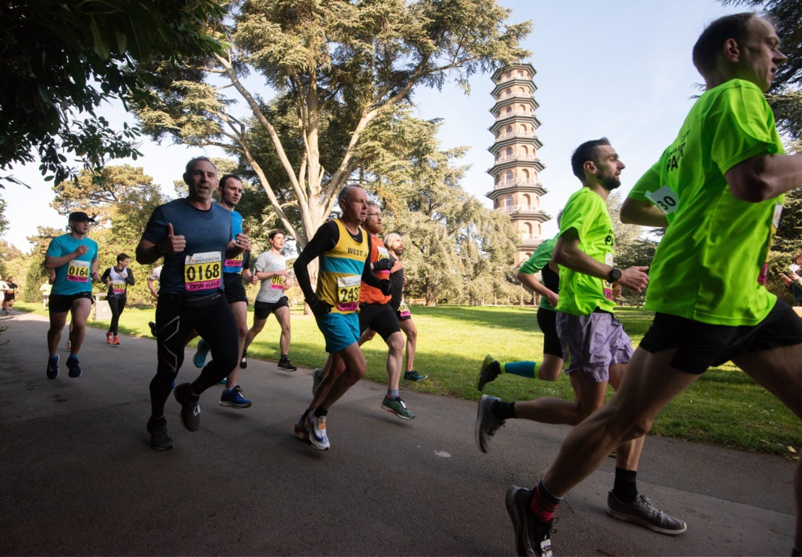 Runners participating in a race on a sunny day, passing a tall pagoda in the background. The runners are dressed in colorful athletic gear, and some wear bib numbers. The scene is set in a park with trees and well-maintained paths.