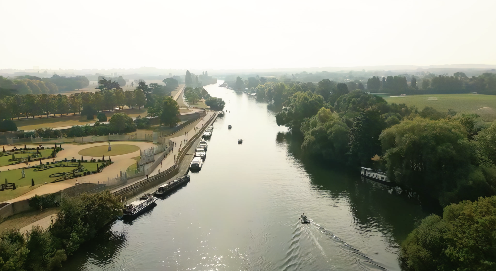 A tranquil river scene with several boats sailing and docked along a tree-lined waterway. On the left, there's a park with pathways and greenery. The horizon is faintly visible under a hazy sky, with a soft focus on distant trees and landscape.