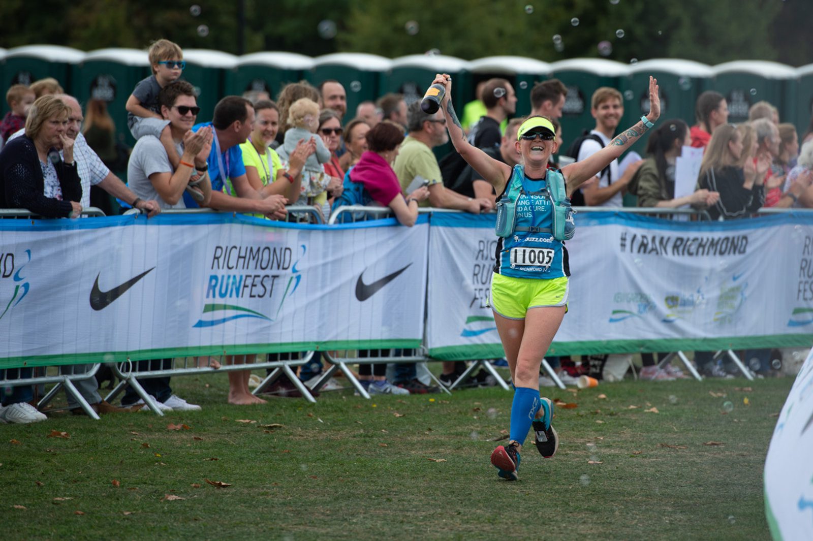 A joyful runner wearing a bright neon outfit and bib number 11005 celebrates while crossing the finish line at the Richmond Runfest. Spectators cheer from behind barriers with event signage. Other runners and portable toilets are visible in the background.