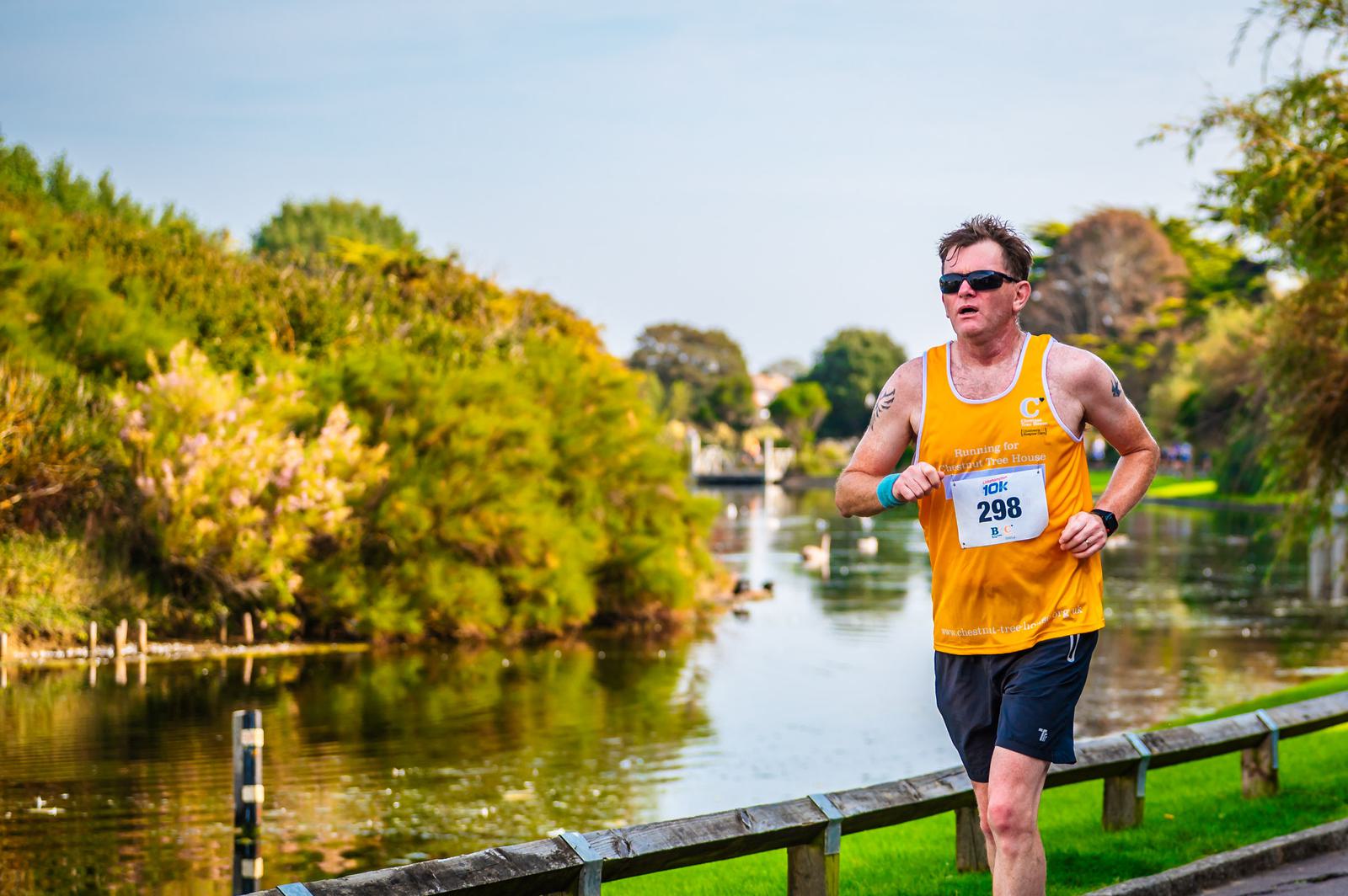 A man wearing a yellow tank top and black shorts, running along a pathway beside a peaceful river, surrounded by green trees and bushes. He is wearing sunglasses and has a race number 298 pinned to his shirt. The sky is clear and sunny.