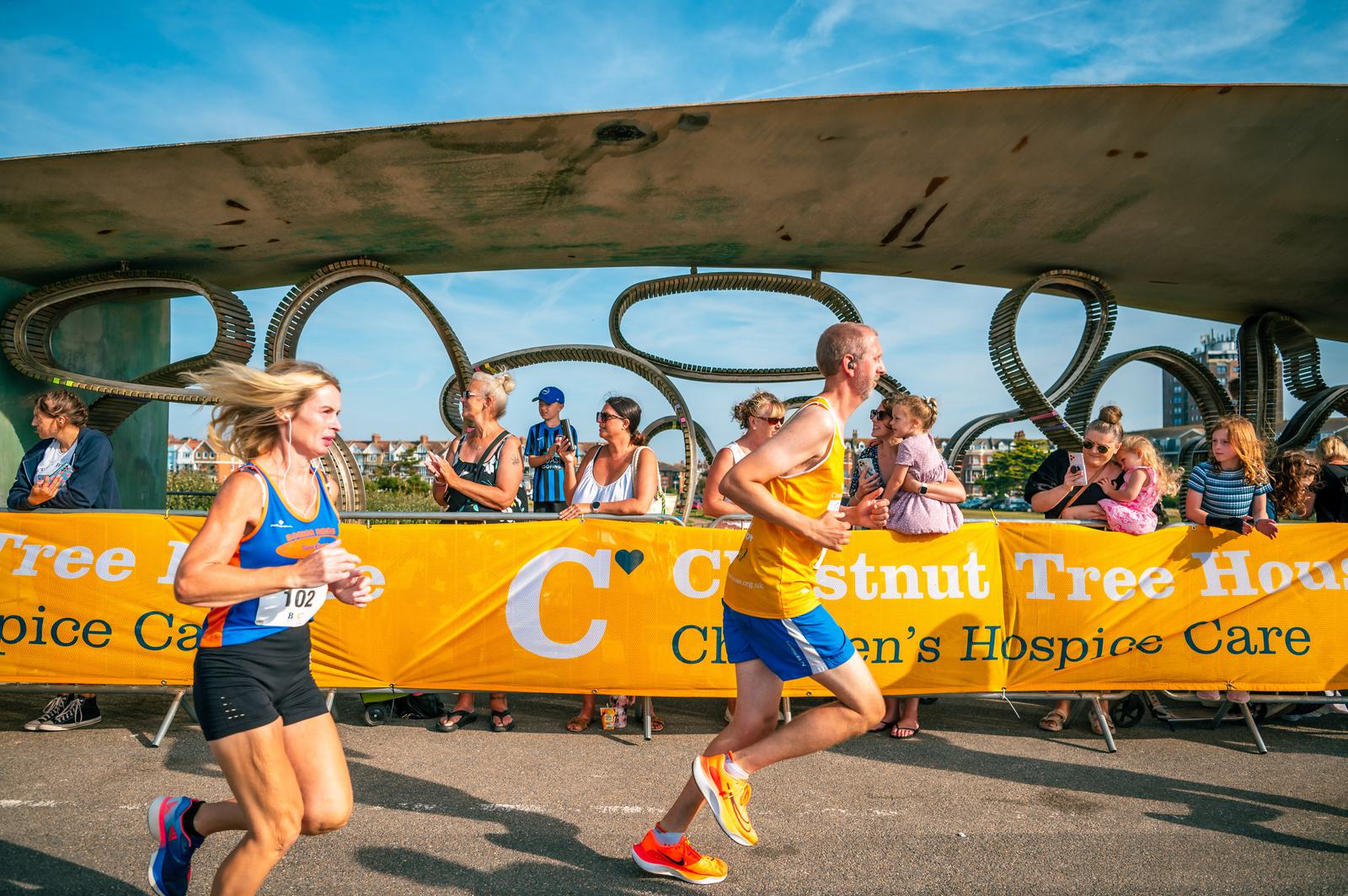 Two runners participate in a race, passing a crowd of spectators. A yellow banner for a children's hospice care event is visible in the background. The sky is clear and sunny.