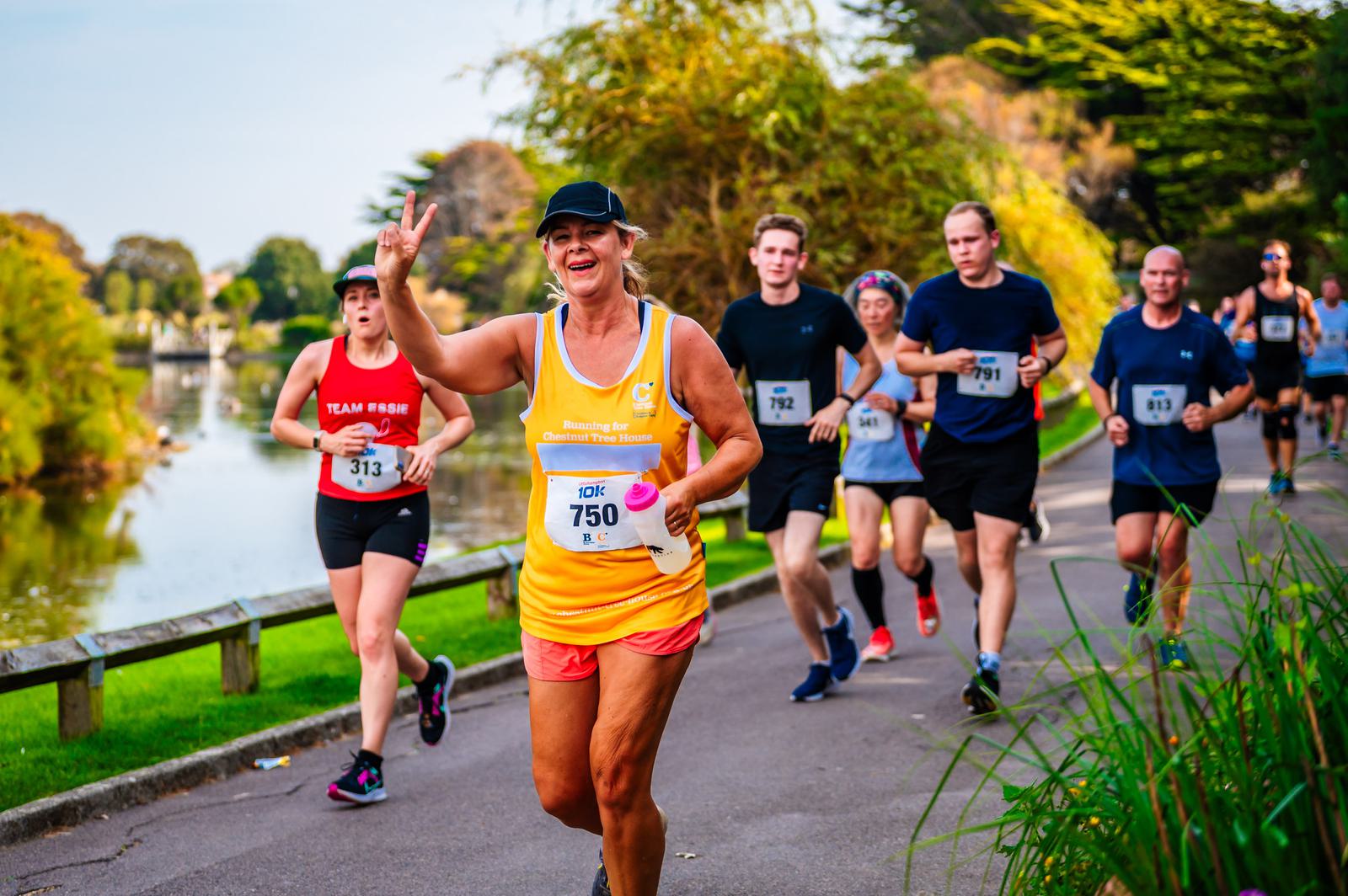 A group of runners participating in a marathon. The runner in the foreground, wearing a yellow tank top and black cap, is smiling and making a peace sign. They are running on a path beside a river, surrounded by greenery. Other runners are visible behind.