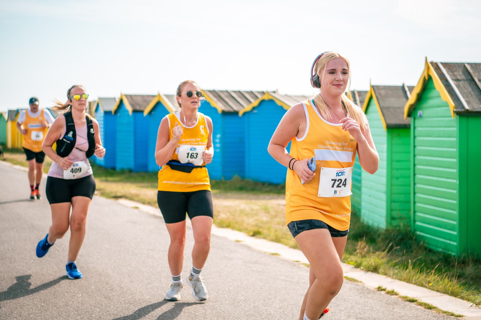 Three women in yellow shirts and running gear jog along a path beside colorful beach huts. They have race numbers pinned to their shirts. It’s a sunny day with blue skies.