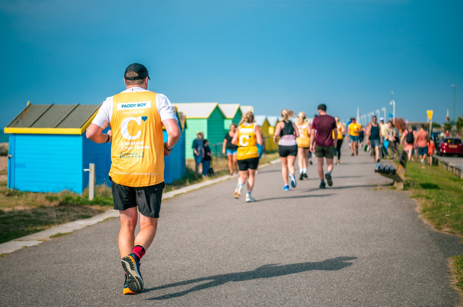 People participating in a charity run on a sunny day, jogging along a paved coastal path. They wear yellow vests with numbers. Colorful beach huts line the path, and more runners are visible in the distance.