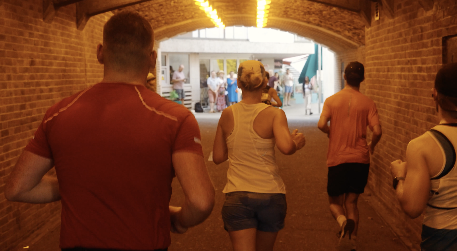 A group of people jog through a brick tunnel illuminated by yellow lights, heading towards an open area with a crowd of onlookers and some buildings in the background. The joggers wear athletic clothing and the tunnel’s brick walls curve overhead.