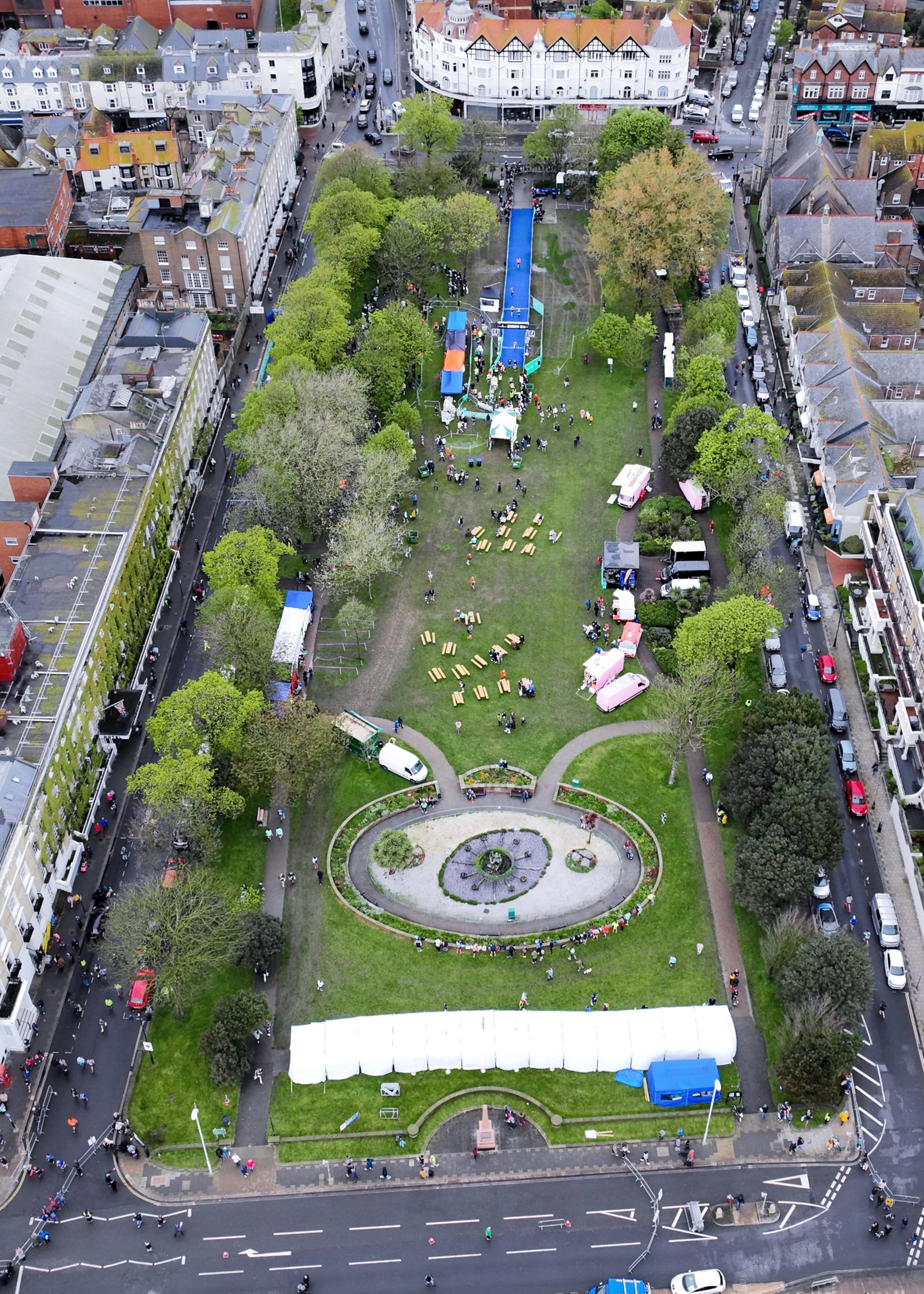 Aerial view of a large, rectangular park with green lawns, trees, tents, and seating areas. People are scattered across the park, participating in various activities. Buildings and streets border the park on all sides.