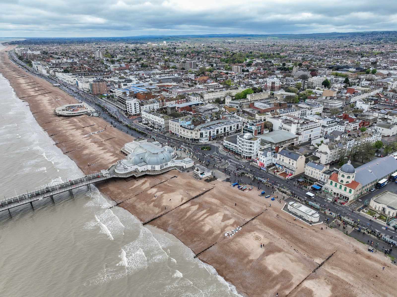 Aerial view of a coastal town featuring a long pier stretching into the sea and a sandy beach with waves coming ashore. Buildings, roads, and greenery make up the busy town, while a partly cloudy sky looms overhead. The town extends inland with numerous structures visible.