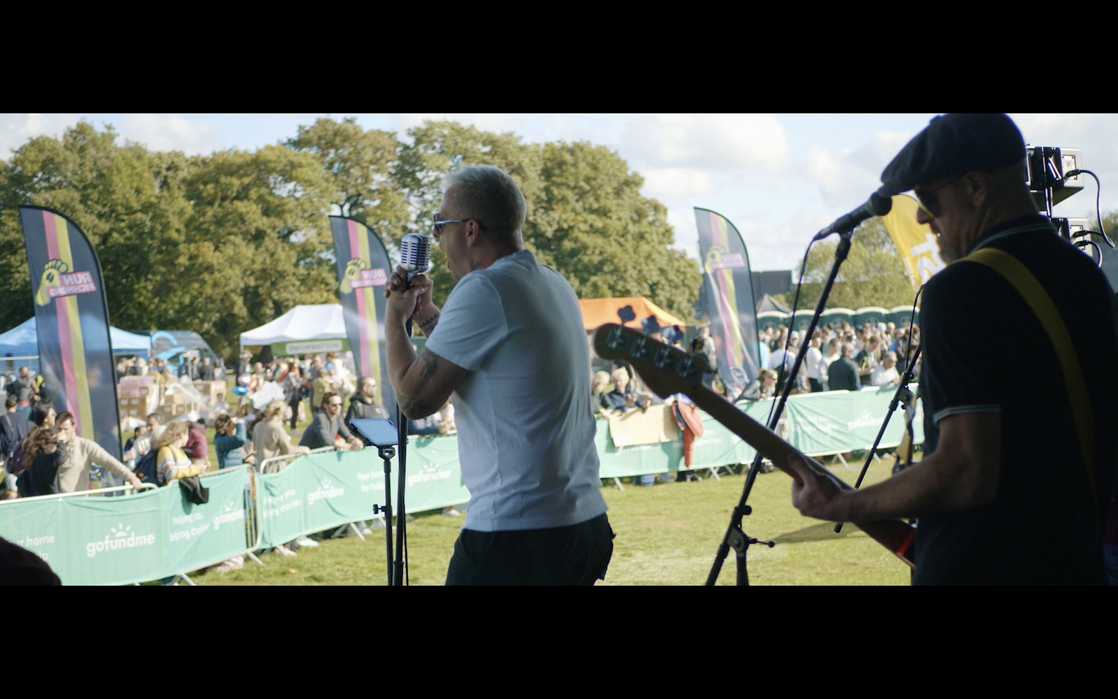 A band performs at an outdoor event. The scene captures a vocalist singing into a microphone and a guitarist playing beside him. The audience is visible in the background, along with event banners, tents, and a sunny sky with trees framing the area.