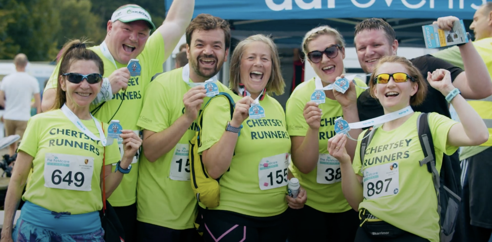 A group of seven smiling runners wearing bright yellow "Chertsey Runners" shirts proudly display their medals. They stand under a blue tent, celebrating their achievement. Each runner has a bib number pinned to their shirt, and some hold drinks in hand.