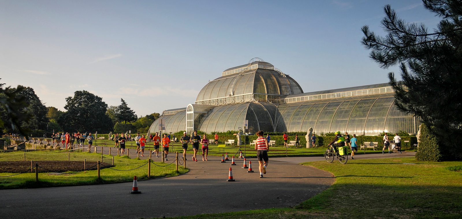 A large group of people, including runners and cyclists, gather in front of a large, ornate greenhouse on a sunny day. The greenhouse, made of glass and iron, stands prominently in the background with well-maintained lawns and trees surrounding it. Traffic cones mark a pathway.