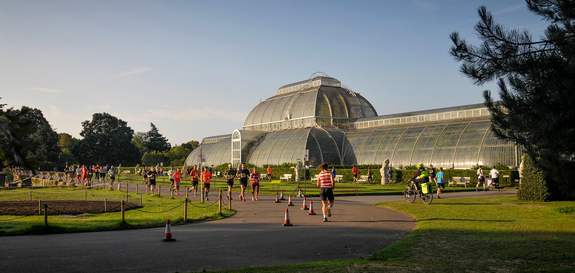 A large group of people, including runners and cyclists, gather in front of a large, ornate greenhouse on a sunny day. The greenhouse, made of glass and iron, stands prominently in the background with well-maintained lawns and trees surrounding it. Traffic cones mark a pathway.