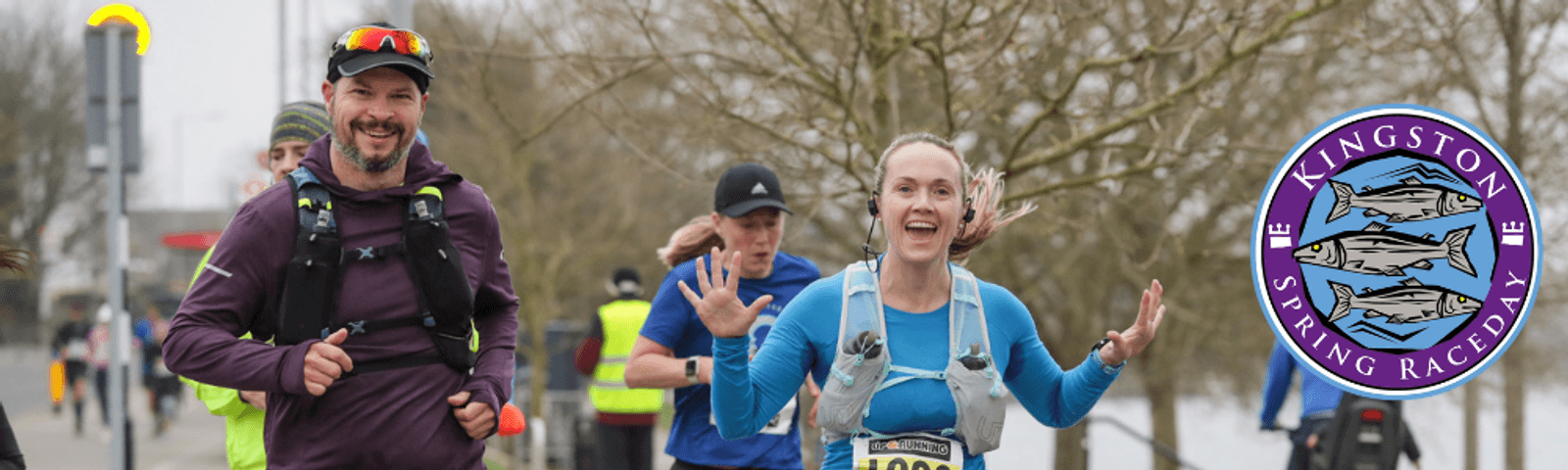 Two smiling runners participate in the Kingston Spring Raceday. The woman in the foreground is wearing a blue outfit and the man next to her is dressed in purple. Other runners and leafless trees are visible in the background. The event logo is on the right side.