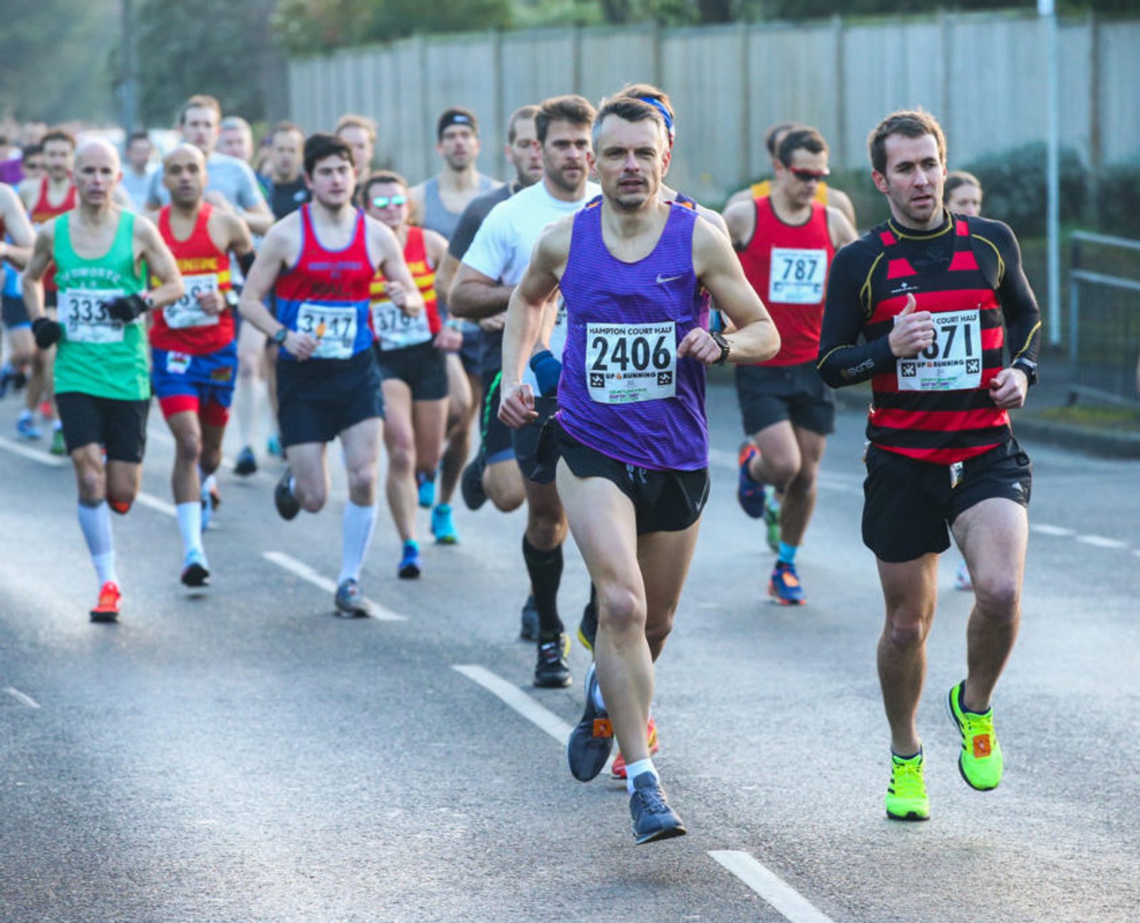 A group of runners in a race, with two men in the forefront wearing numbered bibs 2406 and 1371. The runner in purple is leading, while the runner in red and black is close behind. More runners are visible in the background. The event is taking place on a street.