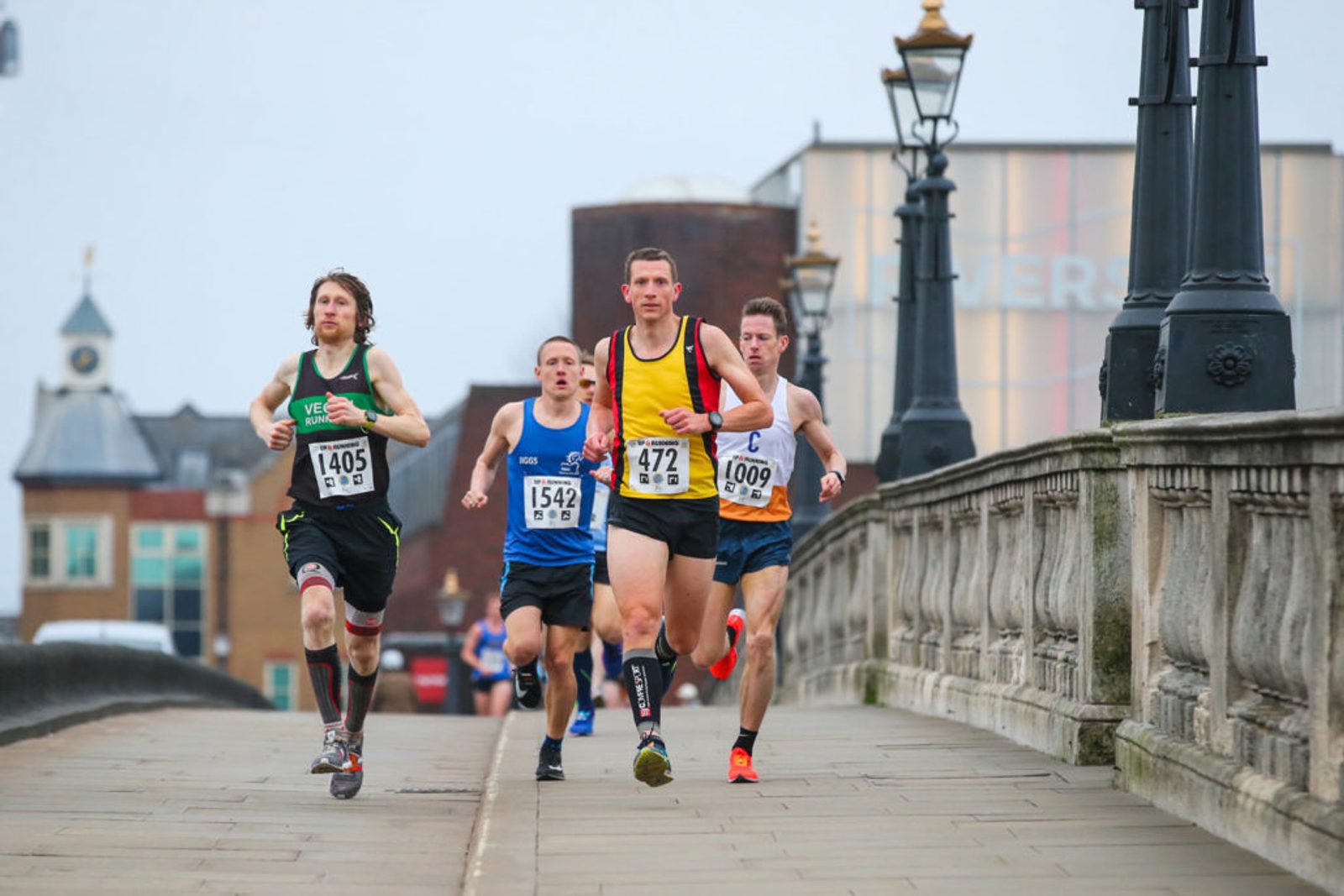 A group of four male runners, all wearing race bibs, are running on a bridge during a race. The runner in front is in a yellow top with bib number 472. Urban buildings and lampposts line the background on a cloudy day.