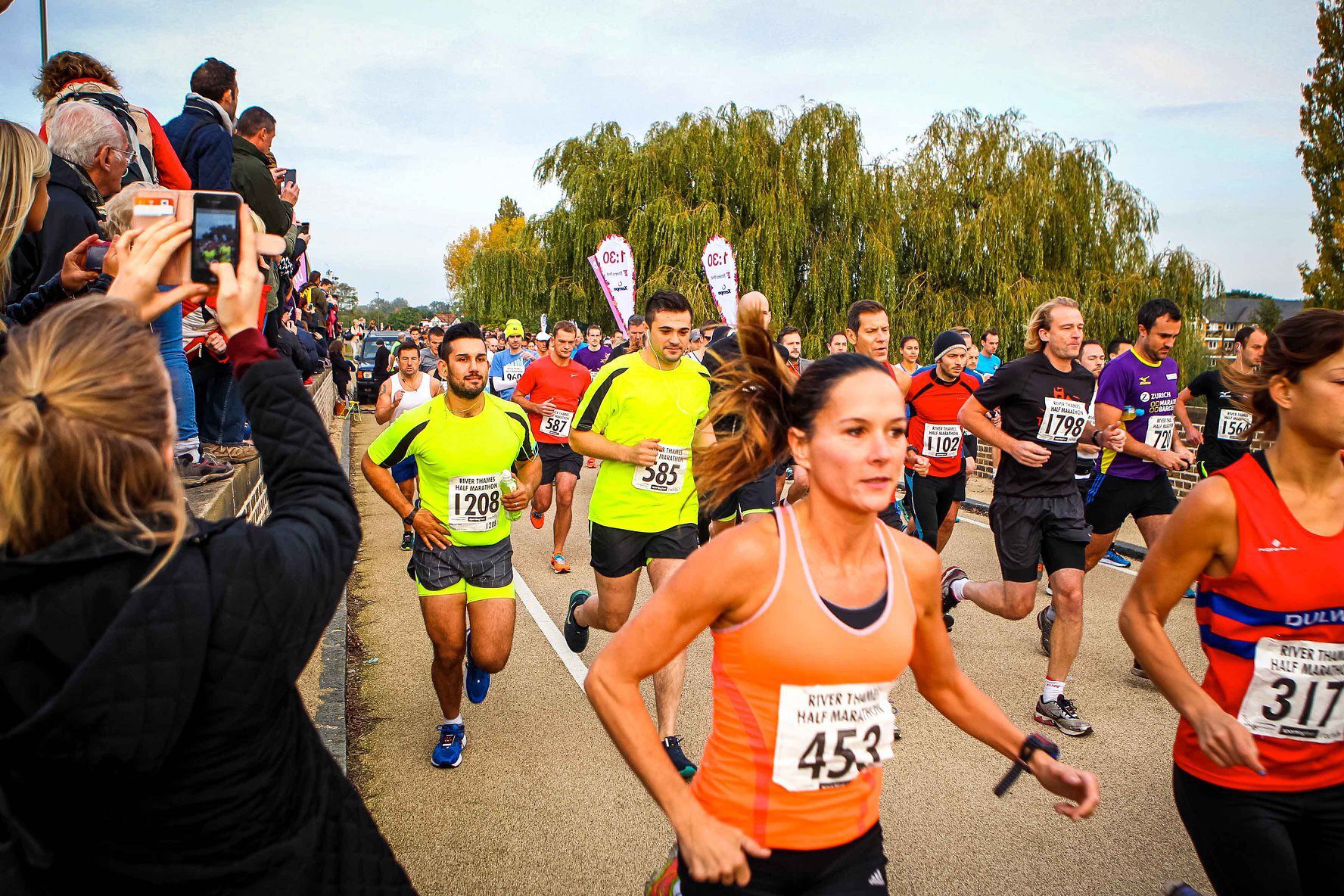 A group of runners participating in a race, some wearing numbered bibs. Spectators and supporters are on the sidelines, cheering and taking photos. The event is taking place outdoors with greenery and large trees in the background. The sky is slightly overcast.
