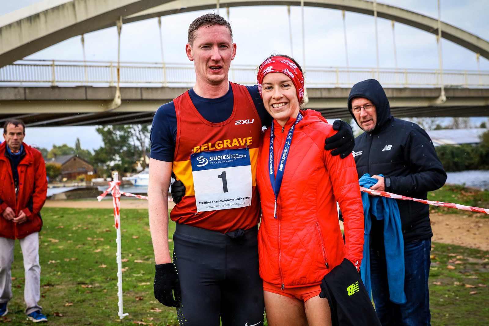 A man and woman, both in athletic gear, smile and pose for a photo after a race. The man wears a racing bib with the number "1" and a "Belgrave" vest, while the woman has a red jacket, headband, and a medal. They stand in front of a bridge, with other people in the background.