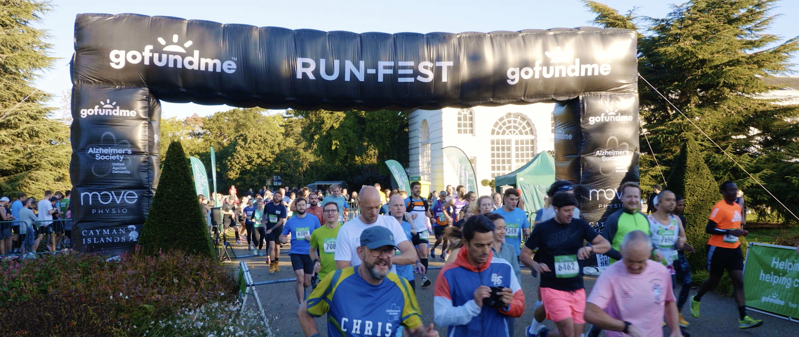 Runners participating in a charity marathon pass under a large inflatable archway labeled "gofundme RUN-FEST." The scene is outdoors, with trees and a building in the background. Participants wear various running attire.