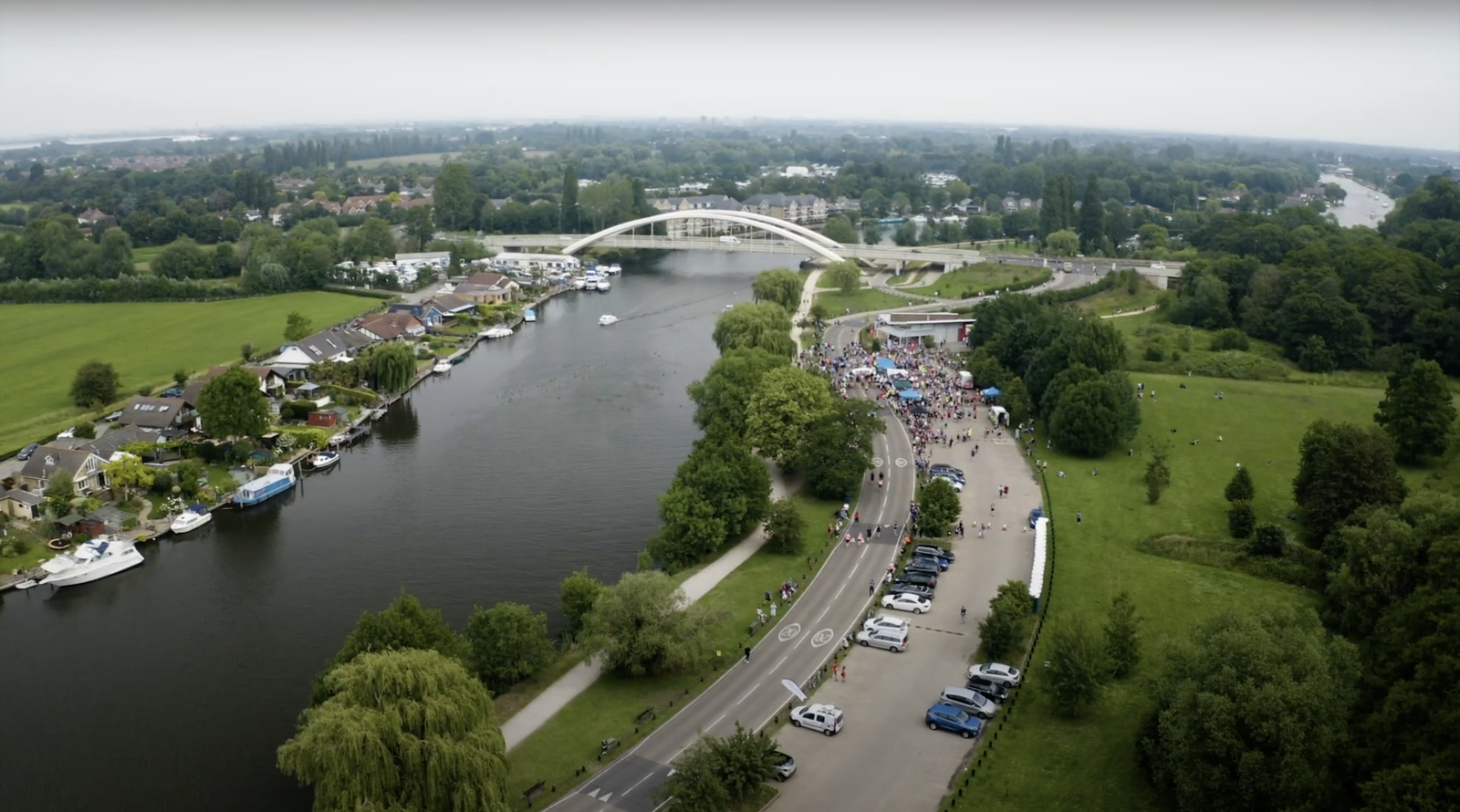 Aerial view of a river with boats, a white arch bridge, and a nearby road with parked cars. A crowd is gathered on the green area by the river. The landscape is lush with trees and greenery, with houses along the riverbank and expansive green fields around.