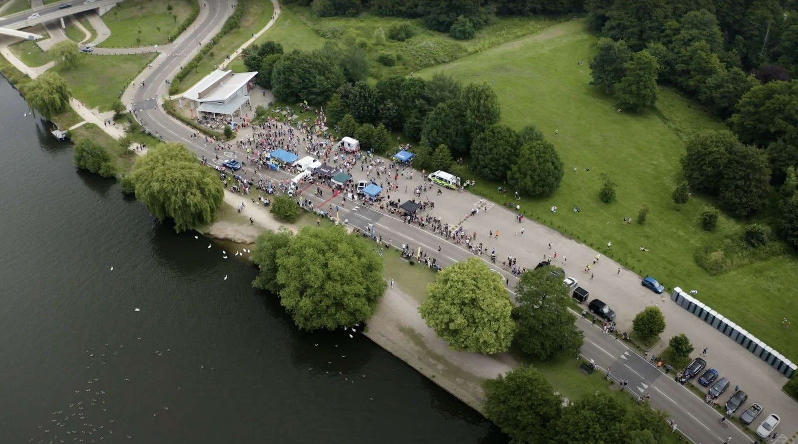 Aerial view of a lakeside park with a road flanked by trees. A large gathering of people is visible near a building. Tents and various colors suggest an outdoor event. The lake is on the left, and adjacent greenery surrounds the scene.