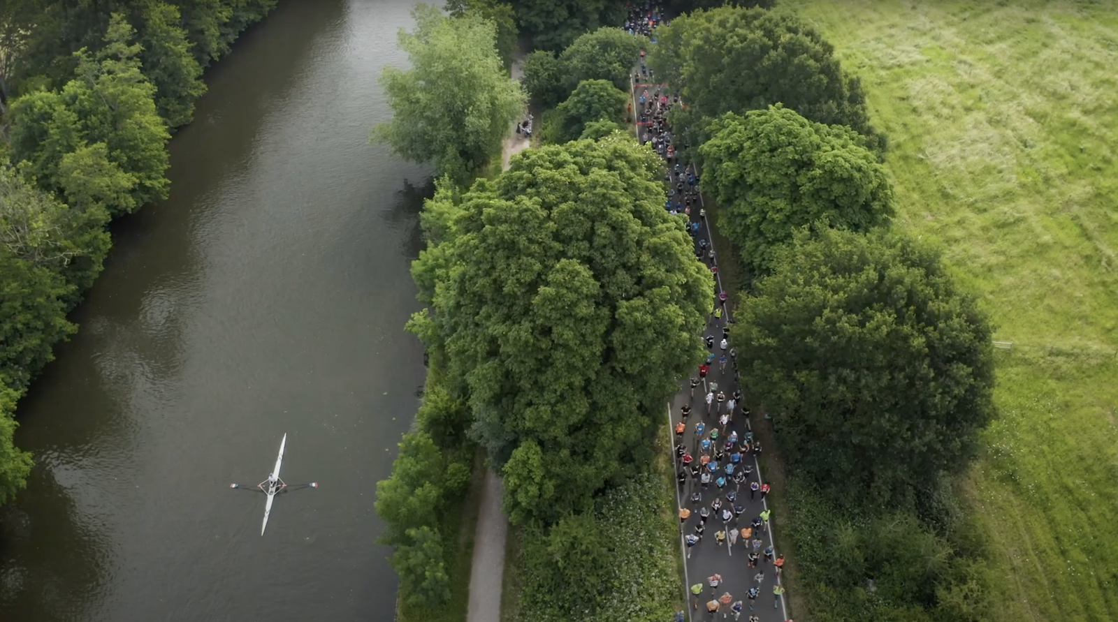 Aerial view of a winding river bordered by lush green trees, with a rower in a boat on the left and a large crowd walking along a path to the right.