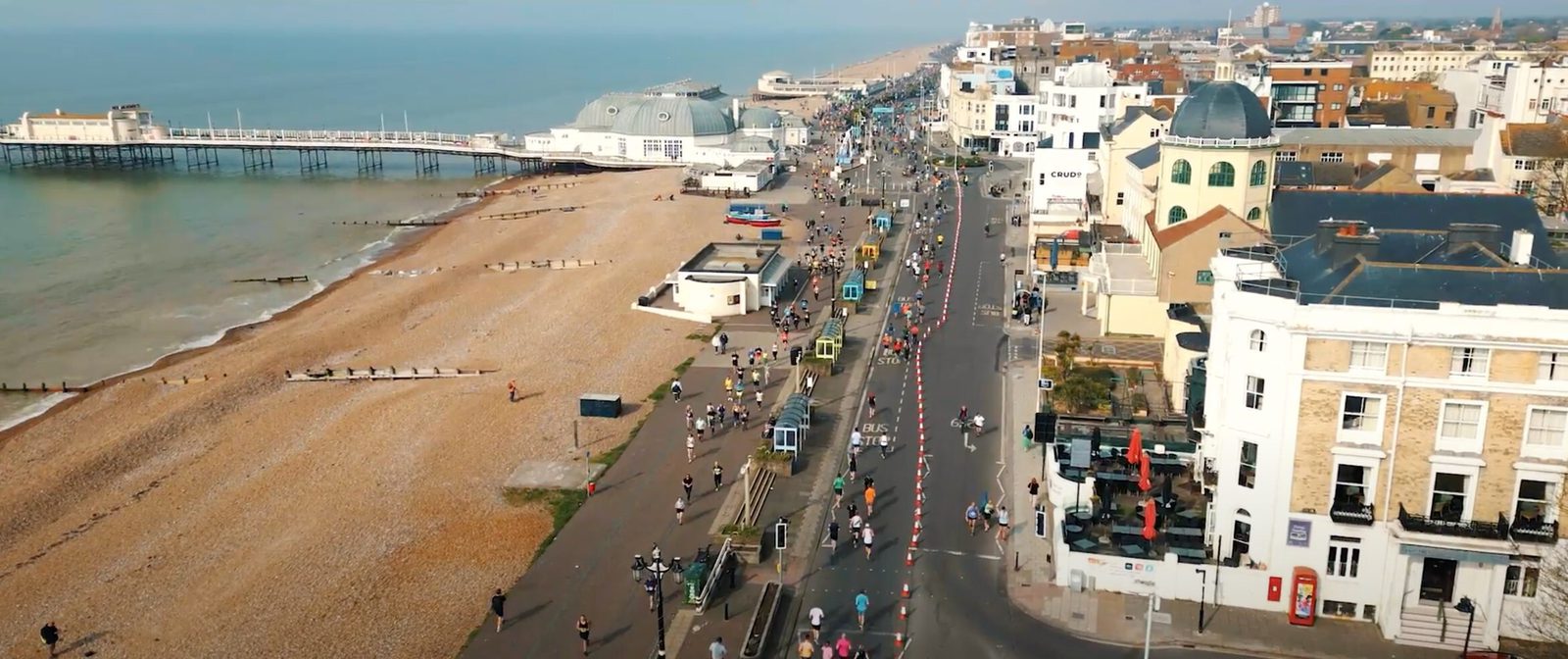 Aerial view of a coastal city with a sandy beach on the left and a city street on the right. People are walking and cycling along the shoreline and the street. Buildings border the street, including shops and housing, with a pier extending into the sea.