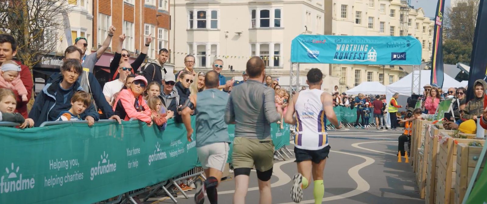 Three runners are approaching the finish line of a street race in an urban area. Spectators line the sides, cheering and taking photos. A blue banner overhead reads "Worthing RunFest" against a backdrop of buildings.