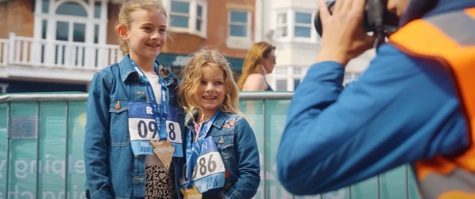 Two young girls wearing blue denim jackets and race bibs with numbers 098 and 086 smile for a photo. They have gold medals around their necks. A photographer in an orange vest takes their picture. There is a background of buildings and a person walking by.