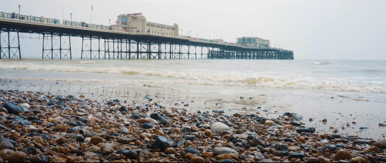 A pebbled beach with wet stones leads to a pier extending over the water. The pier has a building on it, with the sea stretching out into the distance under an overcast sky. Waves gently touch the shore.