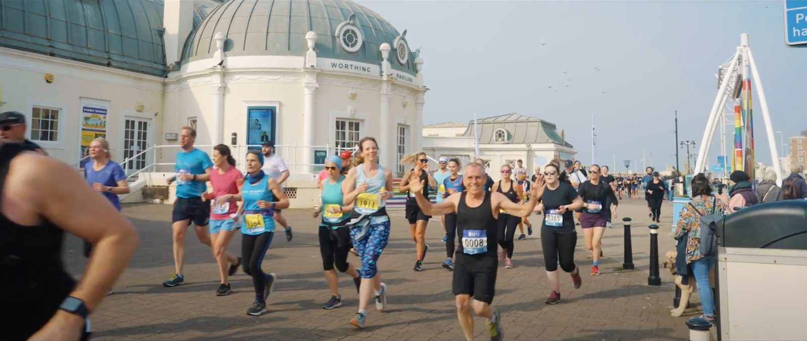 A group of runners participating in a marathon along a coastal promenade with a large domed building in the background. The day appears clear and sunny. Spectators are visible on both sides of the pathway, cheering the runners on.