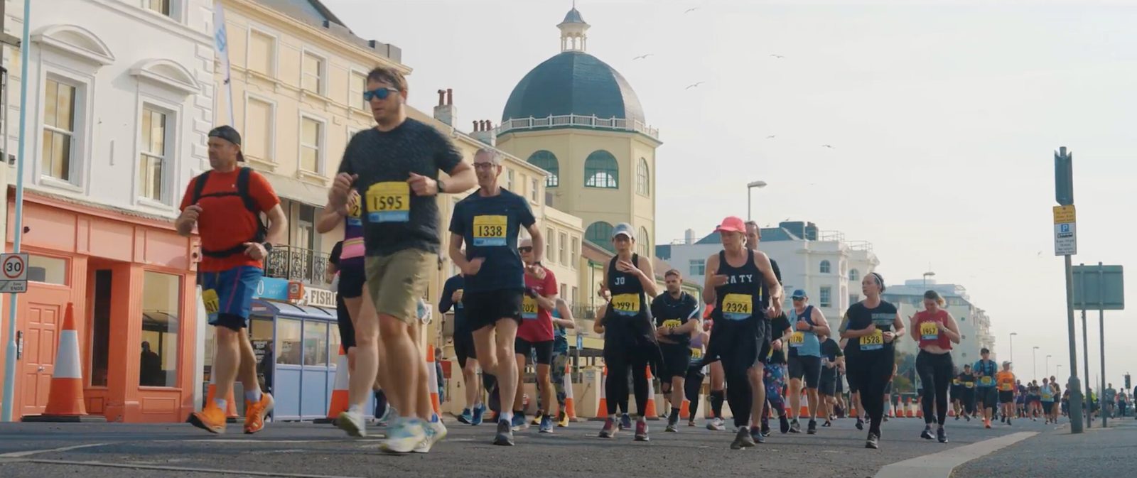 A group of marathon runners jogs down a city street flanked by historic buildings. Runners are wearing numbered bibs and various athletic gear, including hats, sunglasses, and bright sneakers. A domed building stands prominently in the background.