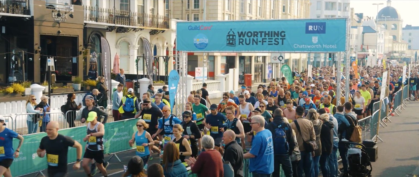 A crowd of runners participating in the Worthing Run-Fest are seen crossing the starting line on a street lined with spectators, signage, and historic buildings. The event banners and volunteers direct and cheer on the participants.