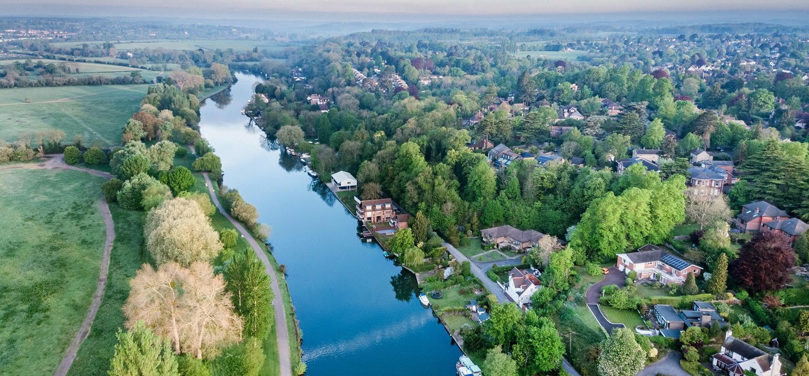 Aerial view of a serene river winding through a lush, green landscape with houses along the banks. The river is surrounded by trees, fields, and residential areas. The sky above is clear with a hint of a misty morning atmosphere.