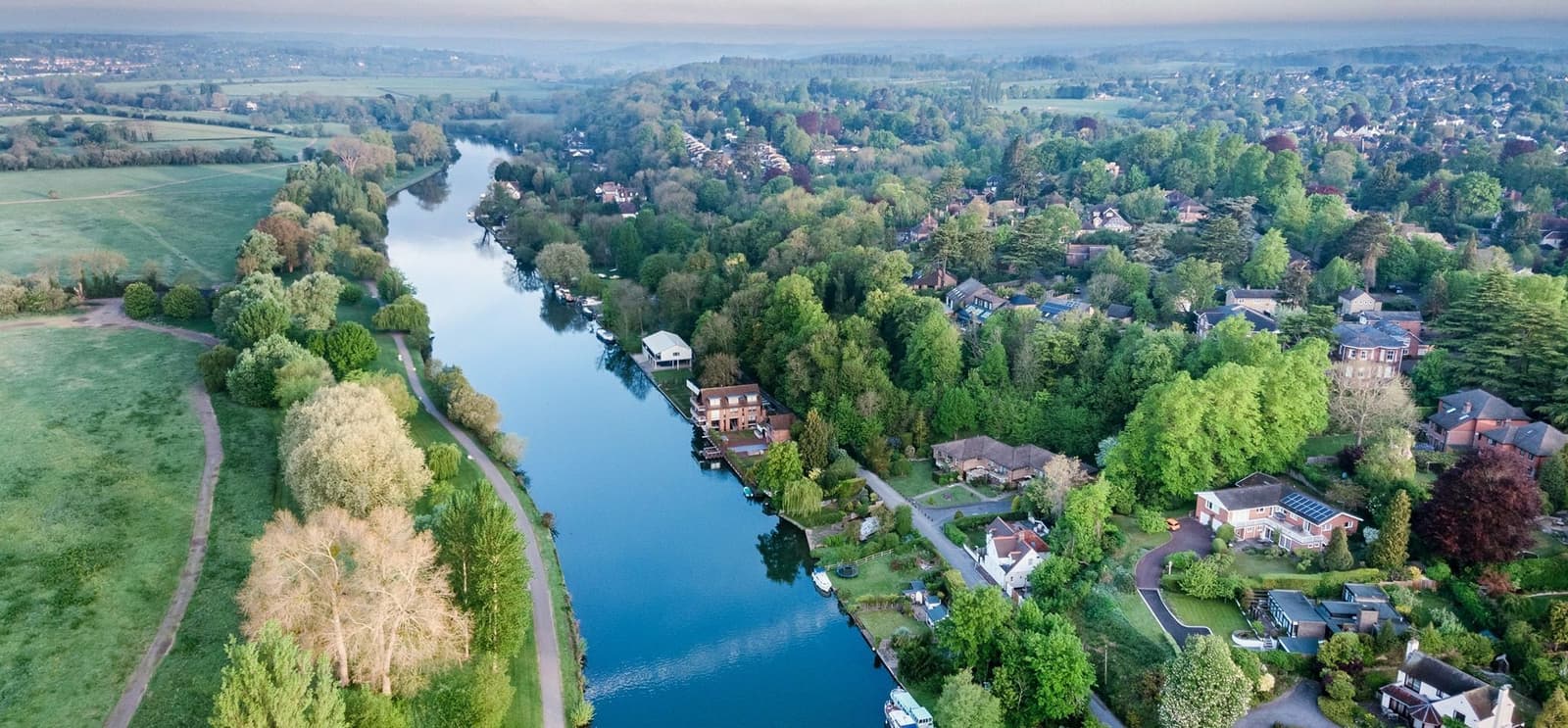 Aerial view of a serene riverside village surrounded by lush greenery. The river winds through the landscape, bordered by trees and quaint houses. The scene includes vast open fields on one side and a mix of residential homes nestled among trees on the other.