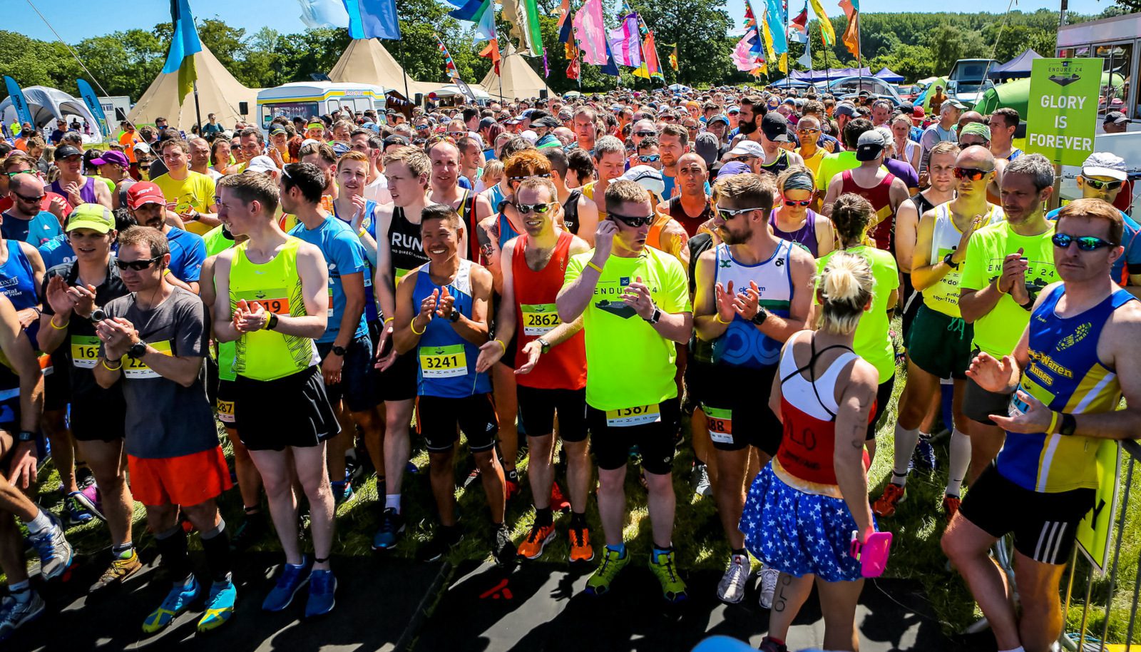 A large group of runners wearing brightly colored athletic wear stands at a starting line, ready for a race. Colorful flags and banners flutter in the background. One participant holds a sign reading “GLORY IS FOREVER.” Some runners clap while others look excited or focused.