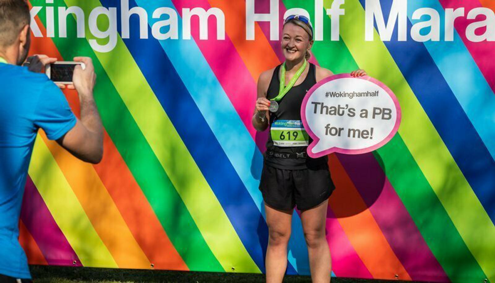 A happy runner posing with a medal in front of a colorful backdrop at the Wokingham Half Marathon. She's holding a speech bubble sign that says, "That's a PB for me!" Another person is taking a photo of her with a smartphone.