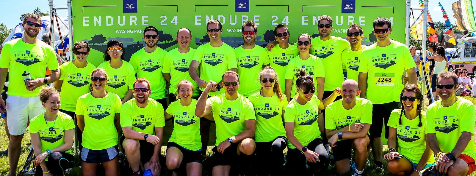 A large group of people wearing matching bright neon green "Endure 24" T-shirts pose for a group photo in front of a green "Endure 24" backdrop at an outdoor event. Some wear sunglasses, and the atmosphere appears to be cheerful and energetic.