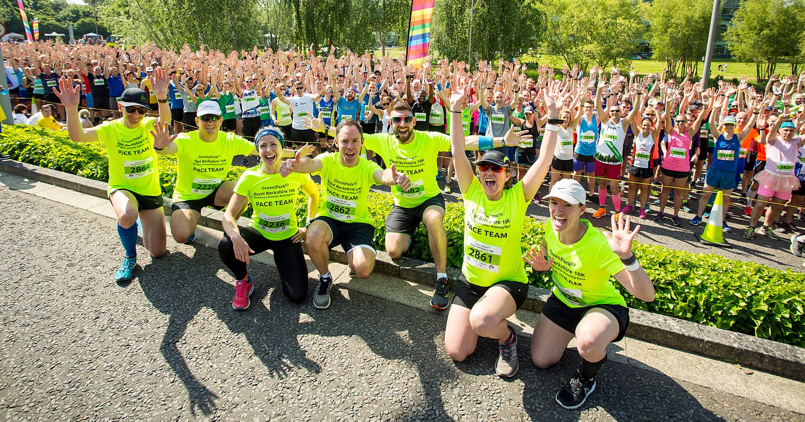 A group of runners wearing bright yellow shirts and bibs pose energetically at the front of a large crowd of participants in a marathon event. The background is filled with a diverse group of runners, all smiling and raising their hands in excitement. Trees and greenery are visible.