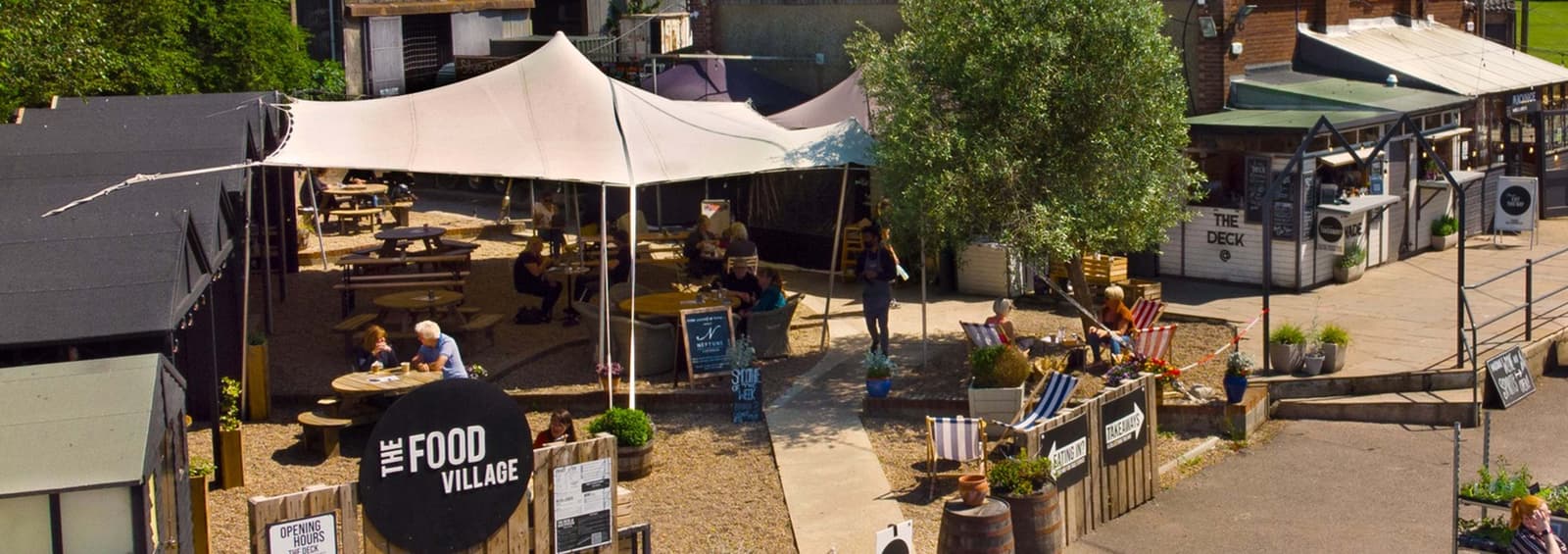 Aerial view of an outdoor food village with multiple seating areas under a white canopy. People are seated at round tables, enjoying food and drinks. Surrounding stalls display signs, and potted plants are scattered around the area.