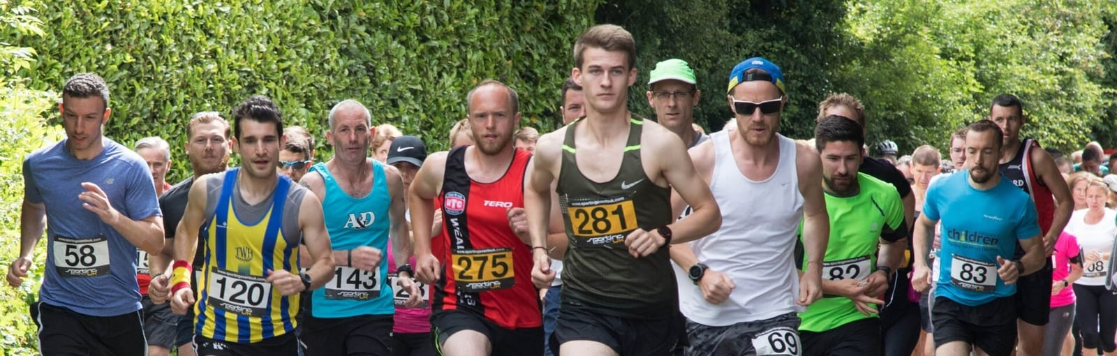 A group of runners, dressed in athletic gear and wearing numbered bibs, compete in an outdoor race on a path surrounded by greenery. The runners vary in age and gender, and the crowd trails closely behind.
